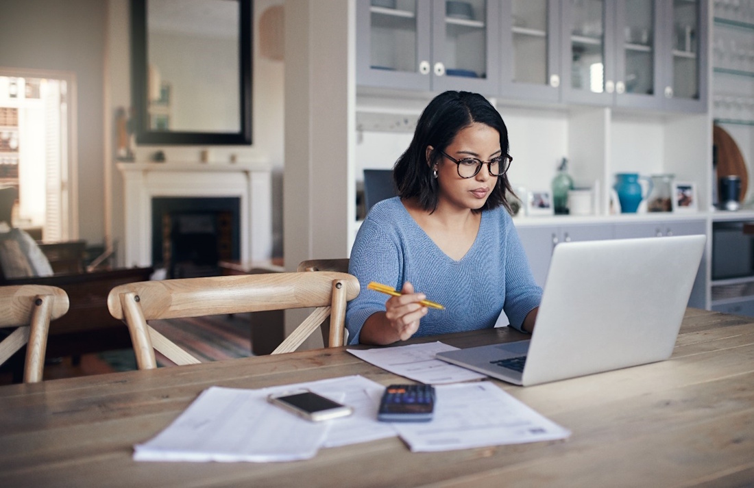A dark-haired woman in a pale long-sleeved shirt and black glasses sits at a wooden table in a large, open kitchen and intently types on a laptop, with various papers, a calculator, and a mobile phone on the table around her.