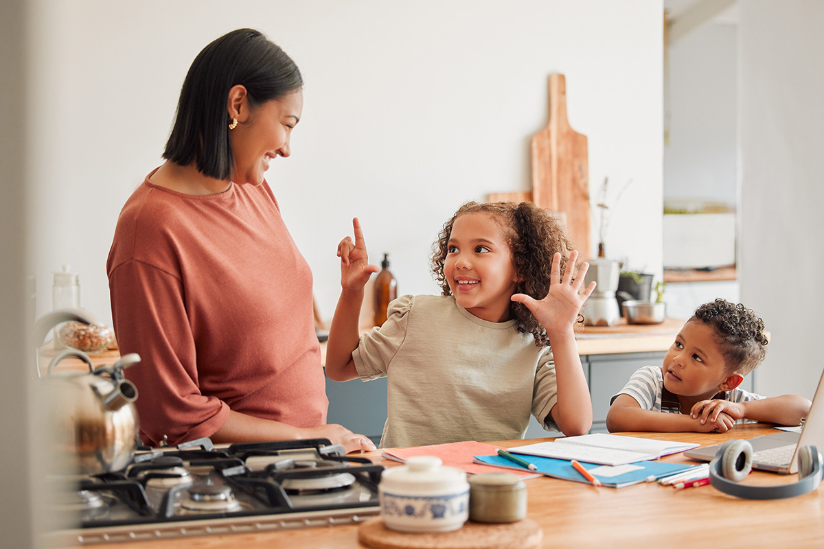 Image Alt Text	A cheerful mother smiles at her two young children at a kitchen counter. The older child excitedly raises her hands while the younger child looks on with curiosity. The counter is covered with school supplies, a laptop, and kitchen items.