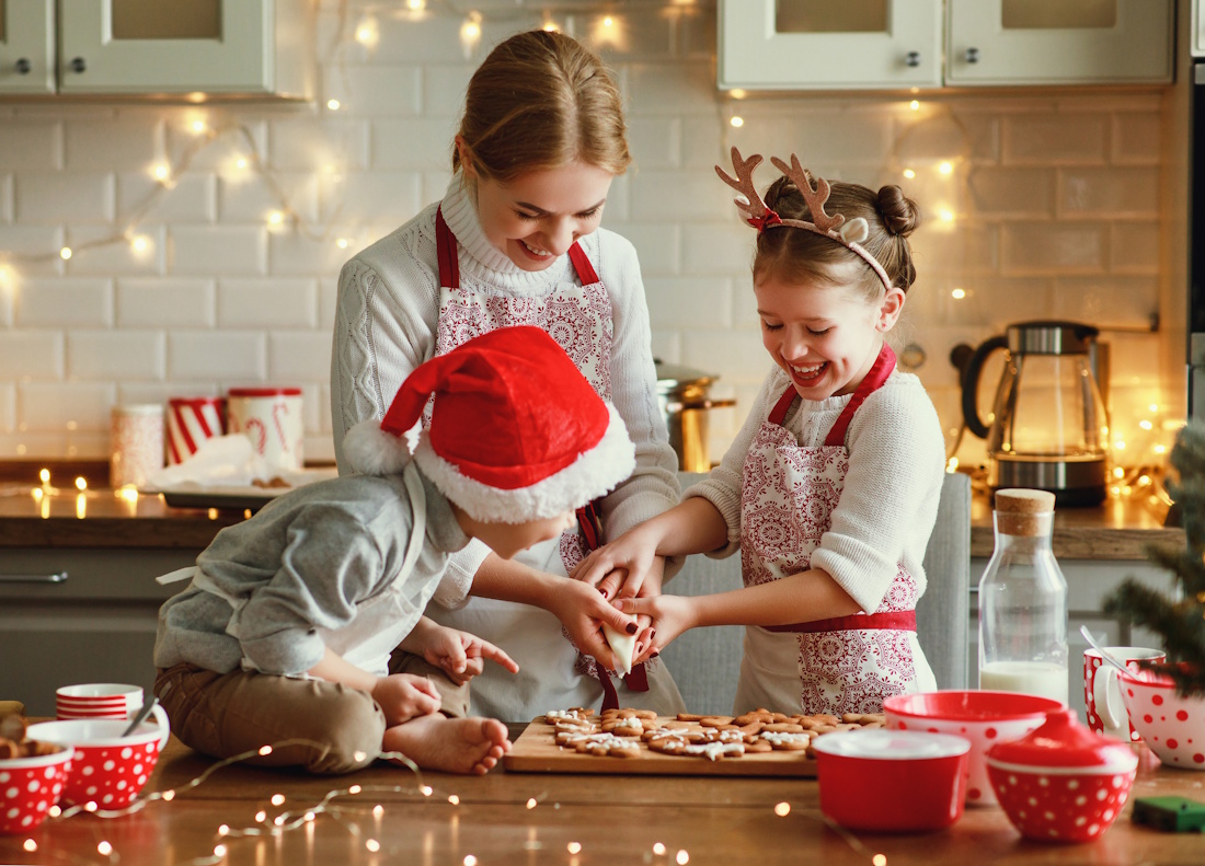 A woman and two children decorate cookies in a kitchen trimmed with holiday lights. The woman and the girl wear matching aprons. The  girl also wears a headband with reindeer antlers. The toddler wears a white apron and a Santa hat.