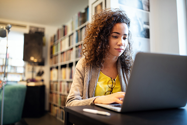Young woman working on laptop in a room with a lot of books on a bookshelf behind her.