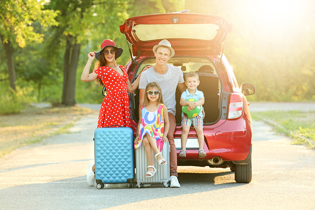 A mom, dad, and two kids behind red car sitting on car, suitcase or standing smiling for a picture.