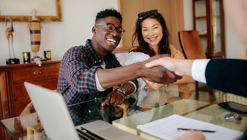 Young couple smiling and shaking hands with someone across a desk or table.