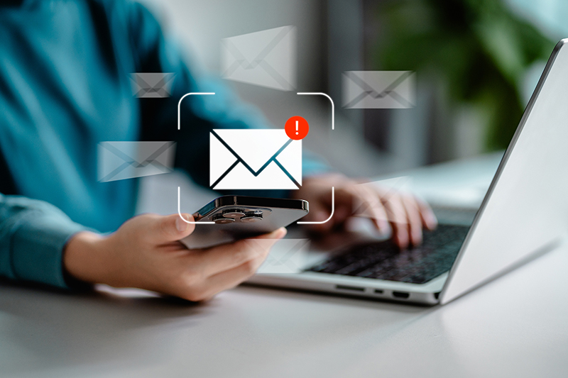 Person sitting at table working on computer and looking at smartphone with an email icon in the center of the screen.