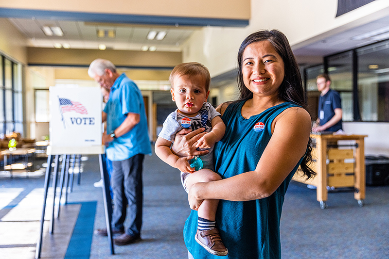 Woman smiling holding toddler son wearing "I Voted" stickers inside of election site.