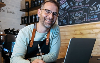 Smiling man wearing glasses and brown apron working on laptop at coffee shop counter with chalkboard menu in background