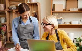 Two women wearing aprons work together at a laptop in a rustic shop with wooden shelves displaying merchandise in the background.