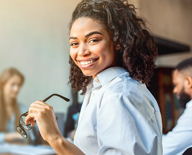 Smiling woman with curly dark hair wearing a light blue shirt, holding sunglasses in an office setting with colleagues in the background