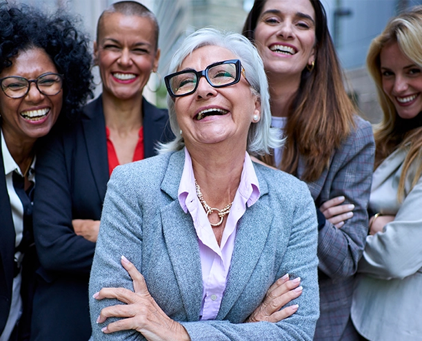 A diverse group of five women in business attire, smiling and looking at the camera. The woman in the foreground is older with gray hair and glasses, laughing.