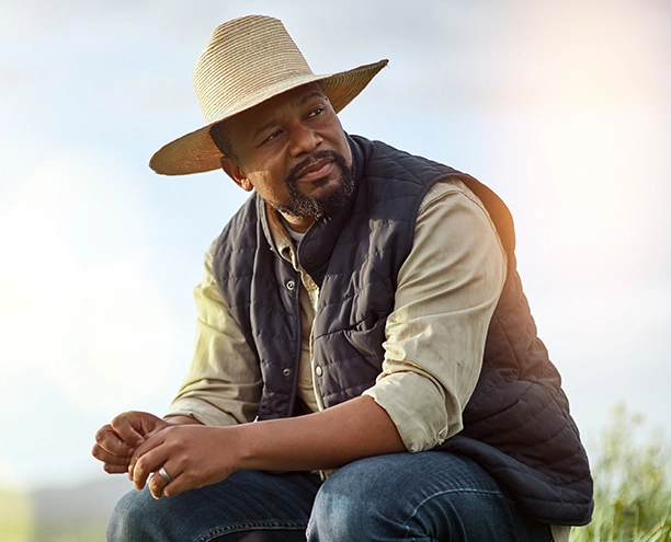 Man wearing a tan cowboy hat, dark vest over light colored shirt, sitting outdoors against a blurred natural background