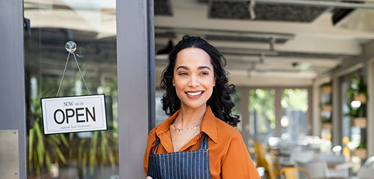 Woman wearing apron standing in front of her store with an "Open" sign on the door.