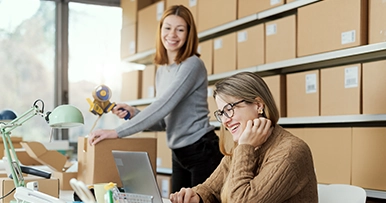 Two women in an office workspace with cardboard boxes on shelves behind them, one standing and one sitting at a desk with a laptop