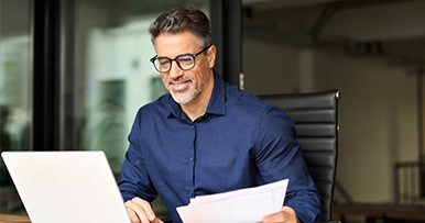 Man in blue shirt wearing glasses working at laptop while holding papers at office desk